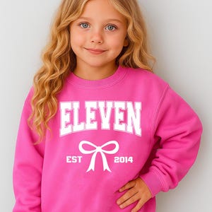 May include: A bright pink sweatshirt with the word "ELEVEN" in bold white letters. Below the word is a white bow graphic, with "EST 2014" printed underneath. The sweatshirt is worn by a young girl with blonde hair.