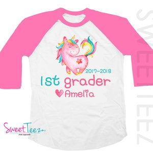 Pode incluir: Camiseta raglan branca e rosa com um design de unic&oacute;rnio em aquarela. A camiseta apresenta um unic&oacute;rnio rosa com uma flor, o texto "1st grader", o nome "Amelia" e o ano "2017-2018". As mangas s&atilde;o rosa brilhante.