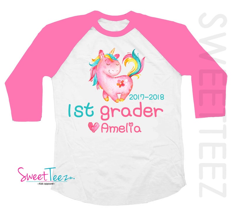 Pode incluir: Camiseta raglan branca e rosa com um design de unic&oacute;rnio em aquarela. A camiseta apresenta um unic&oacute;rnio rosa com uma flor, o texto "1st grader", o nome "Amelia" e o ano "2017-2018". As mangas s&atilde;o rosa brilhante.