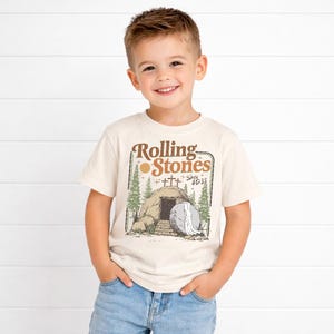 May include: A cream-coloured t-shirt with a graphic that reads "Rolling Stones" above a tomb illustration with a large stone and the text "Since 4033". The shirt is worn by a young child.