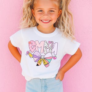 May include: A young girl wearing a white t-shirt with the name "Emma" in a colorful, playful design. The shirt features a bow, pencil, and apple graphic. She has blonde hair and a pink bow in her hair.