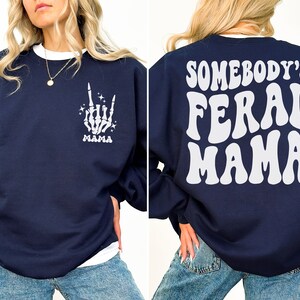 May include: Navy blue sweatshirt with white text and graphics. The front features a skeleton hand with the word "MAMA" below it. The back of the sweatshirt says "SOMEBODY'S FERAL MAMA" in large, white, retro-style lettering. The sweatshirt has a white crew neck.