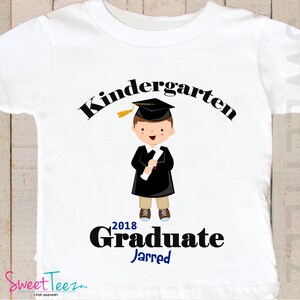 May include: White t-shirt with a black graphic of a boy wearing a graduation cap and gown holding a diploma. The text "Kindergarten Graduate 2018" is above the boy and the name "Jarred" is below the boy.