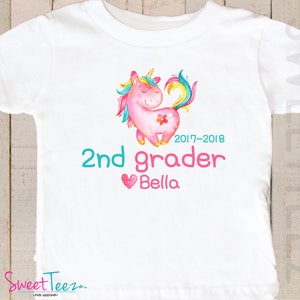 Pode incluir: T-shirt branca com uma ilustra&ccedil;&atilde;o de um unic&oacute;rnio rosa, crina e cauda arco-&iacute;ris. O texto diz "2nd grader Bella" e o ano "2017-2018". A marca "Sweet Teez" est&aacute; na parte inferior. Um design colorido para roupas infantis.