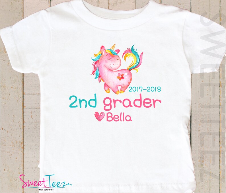 Pode incluir: T-shirt branca com uma ilustra&ccedil;&atilde;o de um unic&oacute;rnio rosa, crina e cauda arco-&iacute;ris. O texto diz "2nd grader Bella" e o ano "2017-2018". A marca "Sweet Teez" est&aacute; na parte inferior. Um design colorido para roupas infantis.