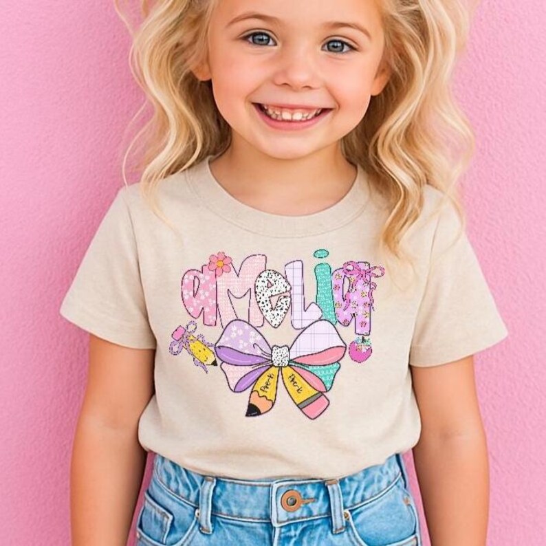 May include: A beige t-shirt with the name "Amelia" in a colorful, whimsical font. The design includes a large bow, pencils, a flower, and a strawberry. The shirt is worn by a young child.