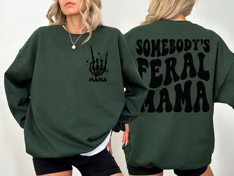 May include: Dark green sweatshirt with the text "SOMEBODY'S FERAL MAMA" on the back. The front features a skeleton hand graphic with the word "MAMA" below. The sweatshirt is a casual, comfortable style.