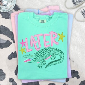 May include: A stack of pastel-colored t-shirts. The top shirt is mint green with the word "LATER" in pink, a green alligator graphic, and yellow star accents. The shirts are stacked on a cowhide patterned surface.