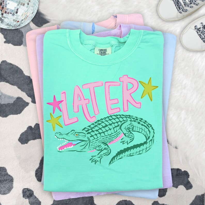 May include: A stack of pastel-colored t-shirts. The top shirt is mint green with the word "LATER" in pink, a green alligator graphic, and yellow star accents. The shirts are stacked on a cowhide patterned surface.