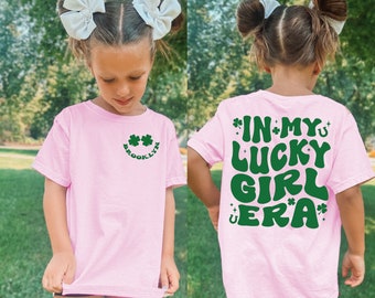 Puede incluir: Camiseta rosa con una carita sonriente de trébol verde en la parte delantera y el texto "In My Lucky Girl Era" en la parte posterior. La camiseta la lleva puesta un niño con lazos blancos en el pelo.
