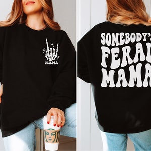 May include: Black sweatshirt with white text and graphics. The front features a skeleton hand with the word "MAMA". The back of the sweatshirt says "SOMEBODY'S FERAL MAMA" in a retro font.