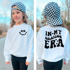 May include: White sweatshirt with black text and graphics. The front features a smiling face with the name "Hadley" and flower accents. The back says "In My Skating Era" with heart accents. The person is wearing a black and white checkered baseball cap.
