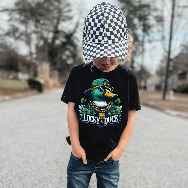 K&ouml;nnte beinhalten: Ein Kind tr&auml;gt ein schwarzes T-Shirt mit einer Comic-Ente, die eine Sonnenbrille und eine goldene Kette tr&auml;gt. Die Ente ist von gr&uuml;nen Kleebl&auml;ttern umgeben und der Text "Lucky Duck" ist zu sehen. Das Kind tr&auml;gt auch eine schwarz-wei&szlig; karierte Baseballkappe und eine blaue Jeans.