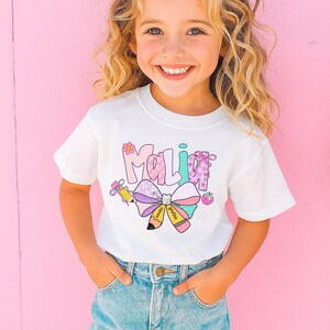 May include: White t-shirt with the name "Malia" in colorful, playful lettering. The design includes a large bow, a pencil, and an apple, all in pastel colors. The shirt is worn by a young girl with blonde hair.