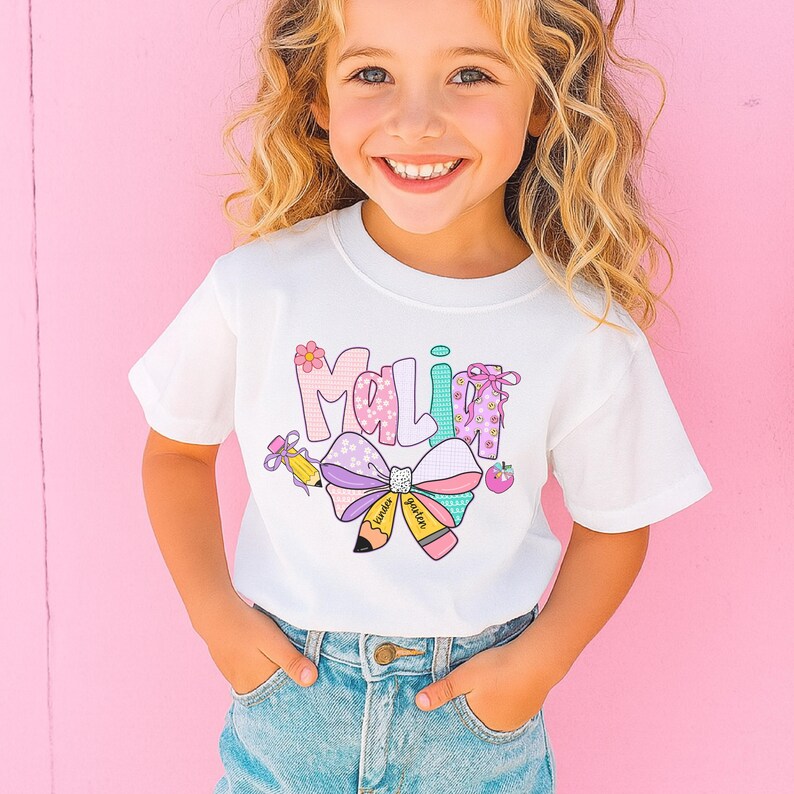 May include: White t-shirt with the name "Malia" in colorful, playful lettering. The design includes a large bow, a pencil, and an apple, all in pastel colors. The shirt is worn by a young girl with blonde hair.
