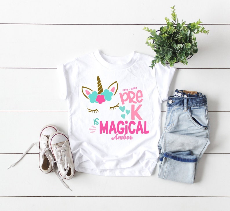 May include: White t-shirt with a unicorn graphic and the text "Pre-K is Magical" in pink and teal. The shirt includes the name "Amber" and the year "2019-2020". A pair of white sneakers and blue jeans are next to the shirt.