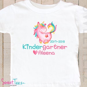 Pode incluir: Camiseta branca com uma ilustra&ccedil;&atilde;o de unic&oacute;rnio rosa e detalhes de arco-&iacute;ris. O texto "Kindergartner Aleena 2017-2018" est&aacute; em turquesa e rosa. Um cora&ccedil;&atilde;o tamb&eacute;m est&aacute; inclu&iacute;do. A camiseta &eacute; para crian&ccedil;as.