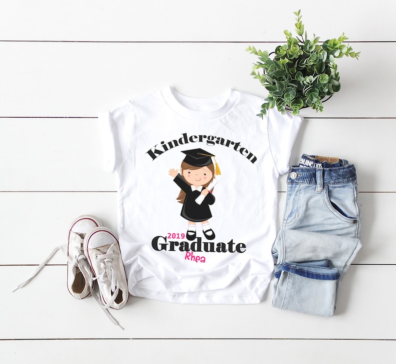 May include: White t-shirt with a black and white illustration of a girl wearing a graduation cap and gown. The text "Kindergarten 2019 Graduate Rhea" is printed on the shirt.