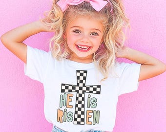 May include: A white t-shirt with a checkered cross and the text "HE is RISEN" in pastel colors. The shirt is being worn by a young girl with blonde hair and a pink bow.