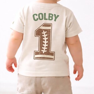 K&ouml;nnte beinhalten: Ein Kleinkind tr&auml;gt ein beiges T-Shirt und khakifarbene Shorts. Auf dem R&uuml;cken des Shirts steht der Name "COLBY" in Gr&uuml;n &uuml;ber einer braunen "1" im Football-Stil. Die Zahl hat wei&szlig;e N&auml;hte.