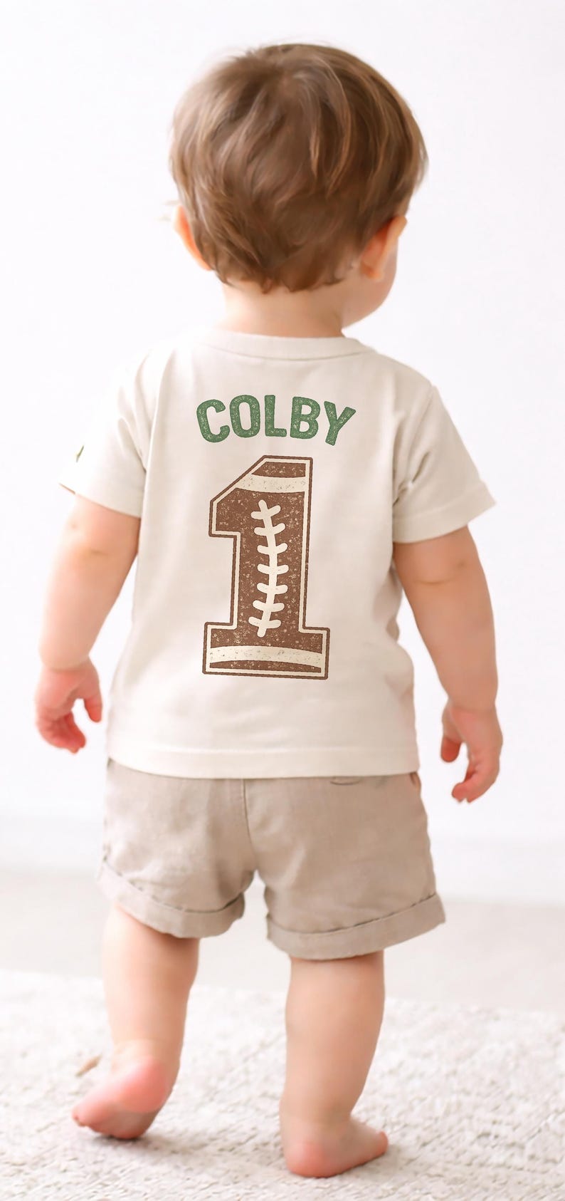 K&ouml;nnte beinhalten: Ein Kleinkind tr&auml;gt ein beiges T-Shirt und khakifarbene Shorts. Auf dem R&uuml;cken des Shirts steht der Name "COLBY" in Gr&uuml;n &uuml;ber einer braunen "1" im Football-Stil. Die Zahl hat wei&szlig;e N&auml;hte.