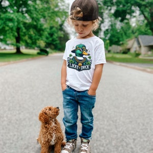K&ouml;nnte beinhalten: Ein kleines Kind tr&auml;gt ein wei&szlig;es T-Shirt mit einer gr&uuml;nen, gelben und schwarzen Comic-Ente, die eine Sonnenbrille und einen Hut tr&auml;gt. Das Shirt sagt "Lucky Duck". Das Kind tr&auml;gt eine blaue Jeans und eine schwarze Baseballkappe. Ein brauner und wei&szlig;er Hund steht neben dem Kind.