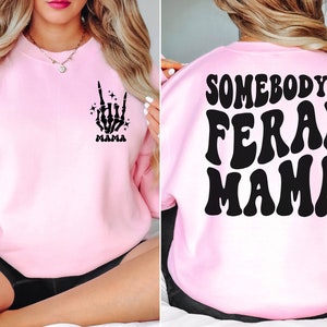 May include: A light pink sweatshirt with a black skeleton hand graphic and the word "MAMA" on the front. The back of the sweatshirt features the text "SOMEBODY'S FERAL MAMA" in a retro font.