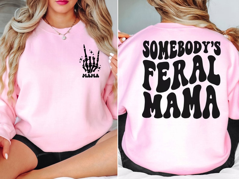 May include: A light pink sweatshirt with a black skeleton hand graphic and the word "MAMA" on the front. The back of the sweatshirt features the text "SOMEBODY'S FERAL MAMA" in a retro font.