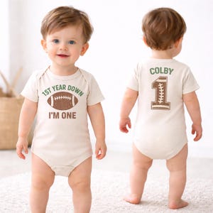K&ouml;nnte beinhalten: Beiger Baby-Body mit Football-Design und dem Text "1st Year Down I'm One" auf der Vorderseite. Auf der R&uuml;ckseite stehen der Name "Colby" und die Zahl "1" im Football-Stil.
