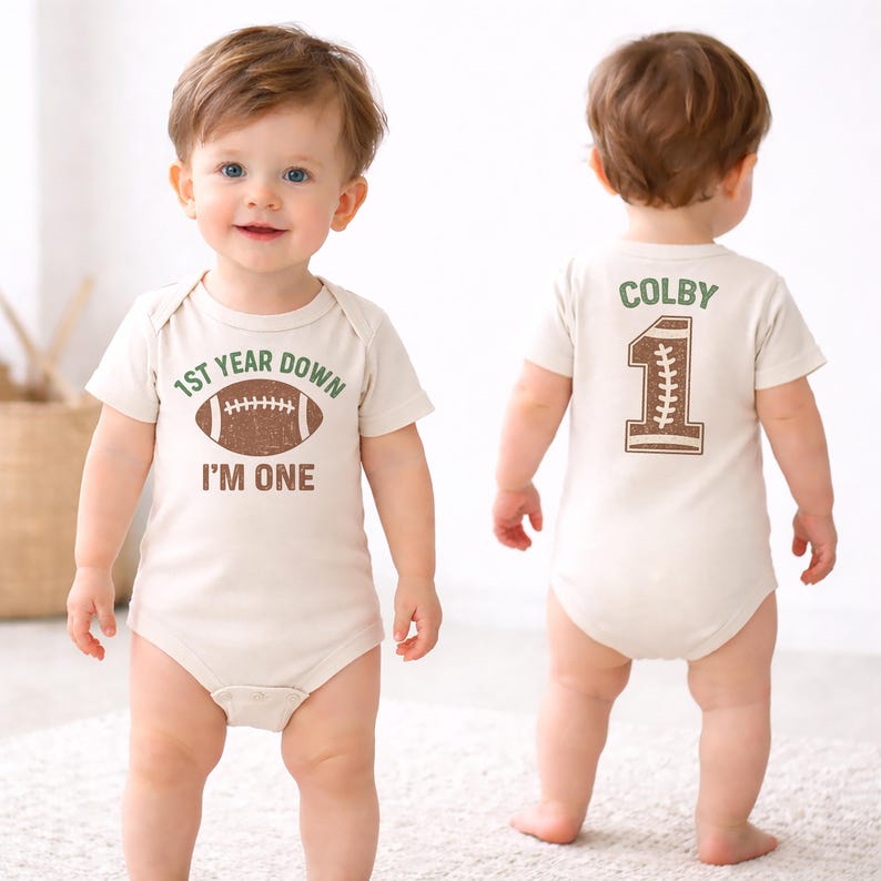 K&ouml;nnte beinhalten: Beiger Baby-Body mit Football-Design und dem Text "1st Year Down I'm One" auf der Vorderseite. Auf der R&uuml;ckseite stehen der Name "Colby" und die Zahl "1" im Football-Stil.