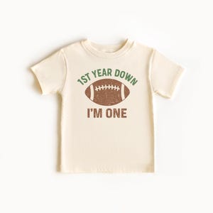 K&ouml;nnte beinhalten: Cremefarbenes kurz&auml;rmeliges T-Shirt mit einem Football-Motiv. Der Text auf dem Shirt lautet "1st Year Down" &uuml;ber einem braunen Football und "I'M ONE" darunter. Das Design ist in Gr&uuml;n und Braun gehalten.