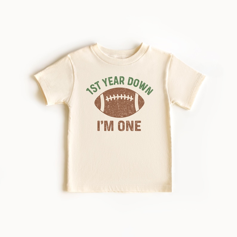 K&ouml;nnte beinhalten: Cremefarbenes kurz&auml;rmeliges T-Shirt mit einem Football-Motiv. Der Text auf dem Shirt lautet "1st Year Down" &uuml;ber einem braunen Football und "I'M ONE" darunter. Das Design ist in Gr&uuml;n und Braun gehalten.
