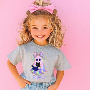 以下が含まれることがあります： ハロウィーンの幽霊のデザインが施されたグレーのTシャツを着た若い女の子。幽霊は本を持っていて、黒猫がいます。シャツには紫色の筆記体で「ソフィア」という名前が書かれています。彼女は髪にピンクのリボンをしています。