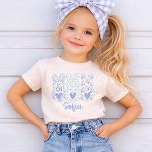 May include: A light beige t-shirt featuring three bunny designs with floral patterns and blue hearts. The name "Sofia" is printed in a script font below the bunnies. The girl is wearing a blue and white gingham headband and denim shorts.