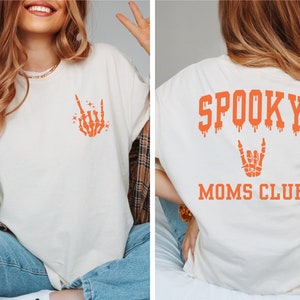 Pu&ograve; includere: Una maglietta bianca con un disegno di mano scheletrica arancione sul davanti e il testo "SPOOKY MOMS CLUB" sul retro.