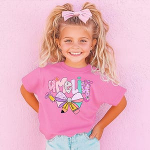 May include: A pink short-sleeved shirt with the name "Amelia" in a colorful, playful font. The shirt features a large bow and pencil graphic, with the words "Second Grade" on the pencil. The girl is wearing blue jeans and a pink bow in her hair.