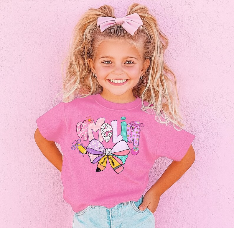 May include: A pink short-sleeved shirt with the name "Amelia" in a colorful, playful font. The shirt features a large bow and pencil graphic, with the words "Second Grade" on the pencil. The girl is wearing blue jeans and a pink bow in her hair.