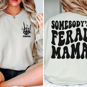 May include: Cream-colored sweatshirt with a black skeleton hand graphic making a rock-on gesture and the word "MAMA" below it. The back of the sweatshirt features the text "SOMEBODY'S FERAL MAMA" in a retro font.