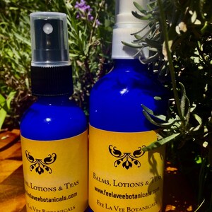 Puede incluir: Dos botellas de vidrio azul con etiquetas doradas y tapas de pulverización negras. Las etiquetas dicen "Balms, Lotions & Teas" y "www.feelaveebotanicals.com" y "Fee La Vee Botanicals".