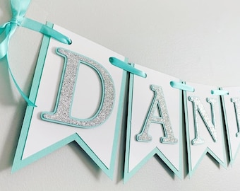 Könnte beinhalten: Ein türkisfarbenes und silbernes Glitzerbanner mit dem Namen "DANIEL" darauf. Das Banner ist aus Karton und hat einen gewellten Rand.