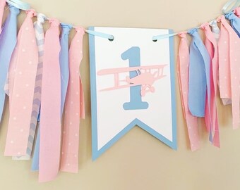 May include: A blue and pink fabric banner with a white card that says "1" and has a pink airplane silhouette. The banner has blue and pink fabric streamers.