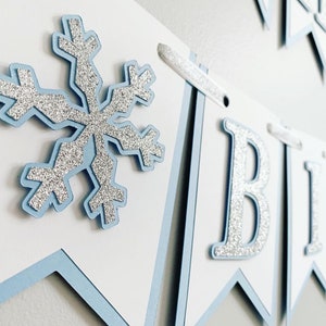 May include: A blue and white banner with silver glitter letters spelling "Happy Birthday". The banner has a snowflake design on one of the flags.