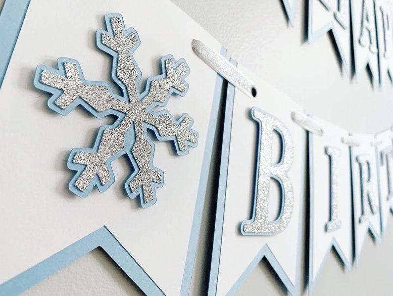 May include: A blue and white banner with silver glitter letters spelling "Happy Birthday". The banner has a snowflake design on one of the flags.