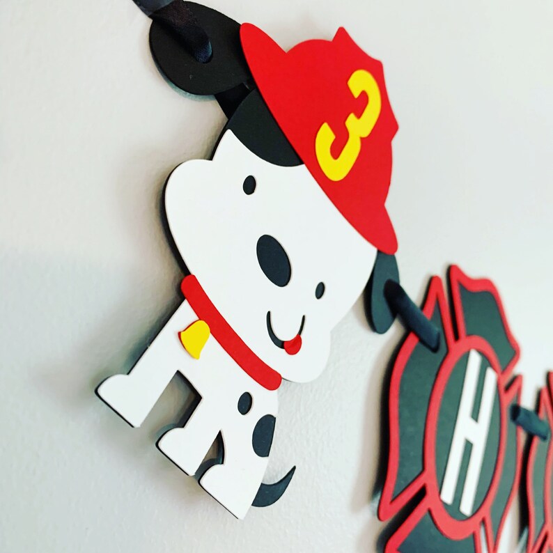 May include: A red, black and white firefighter-themed banner featuring a dalmatian dog wearing a red fireman's hat with a yellow number 3 on it. The banner also includes a black fire hydrant with a white letter H on it.