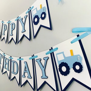 May include: A blue and white banner with the words "Happy Birthday" and two blue tractors. The banner is decorated with blue and white ribbon.