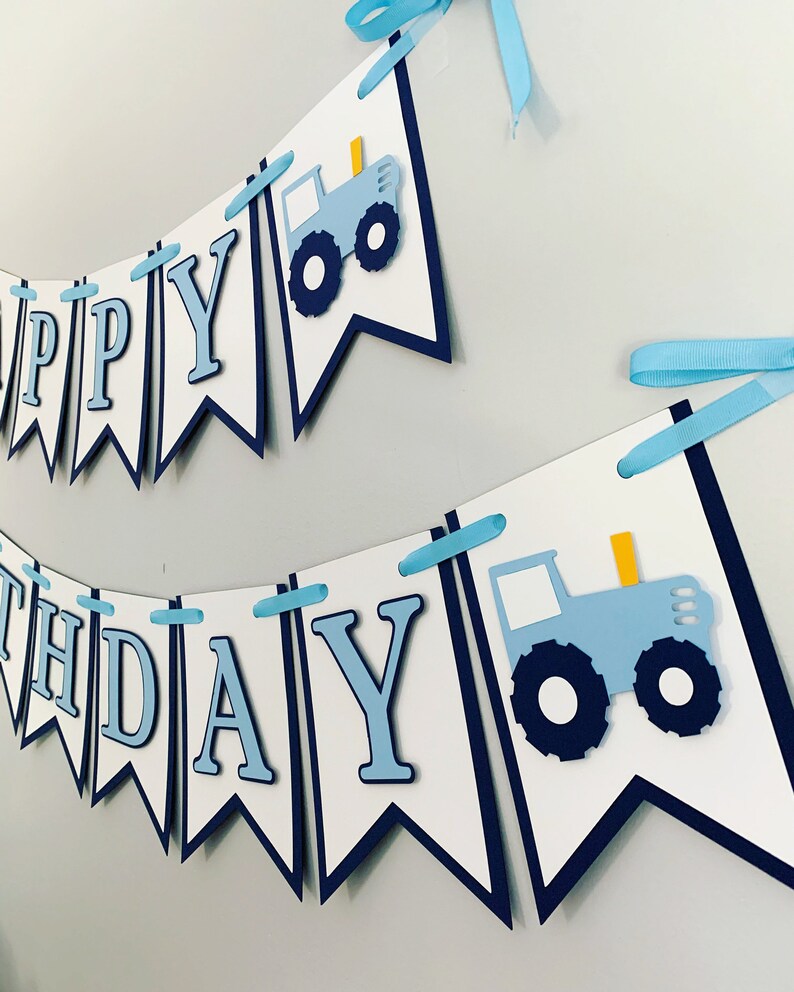 May include: A blue and white banner with the words "Happy Birthday" and two blue tractors. The banner is decorated with blue and white ribbon.