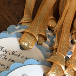 May include: A stack of blue and white baby shower favor tags with gold glitter ribbon. The tags have the text "Baby Jack is coming" and "The time is..." printed on them.