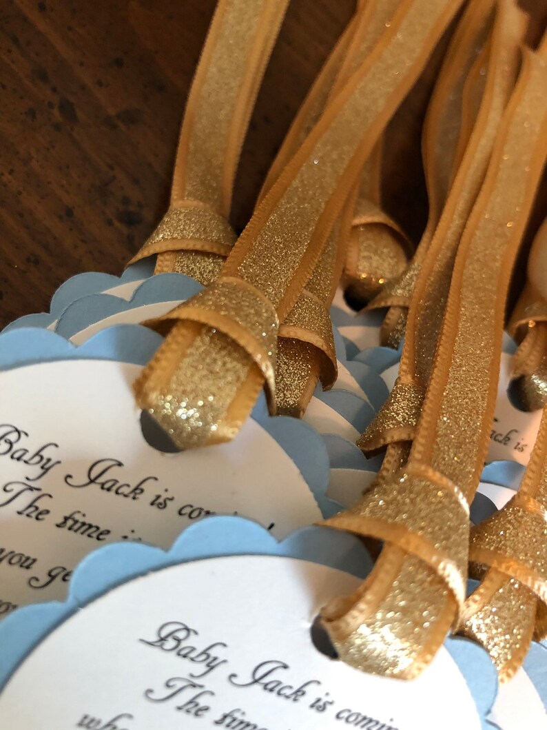 May include: A stack of blue and white baby shower favor tags with gold glitter ribbon. The tags have the text "Baby Jack is coming" and "The time is..." printed on them.