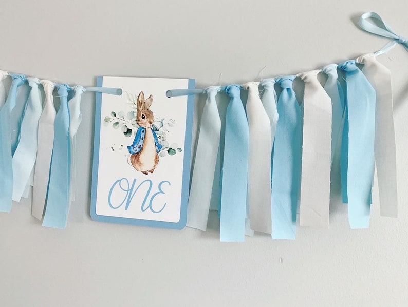Peter Rabbit high chair banner featuring either a blue Peter Rabbit, pink Peter Rabbit, yellow Peter Rabbit or Green Peter Rabbit. The fabric will coordinate with the color of Peter Rabbit's jacket. The banner is available to read an age or name.