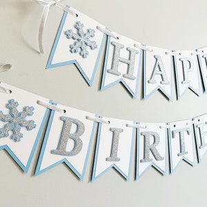 May include: A blue and white banner with silver glitter letters that reads "Happy Birthday". The banner has snowflake cutouts on each end.